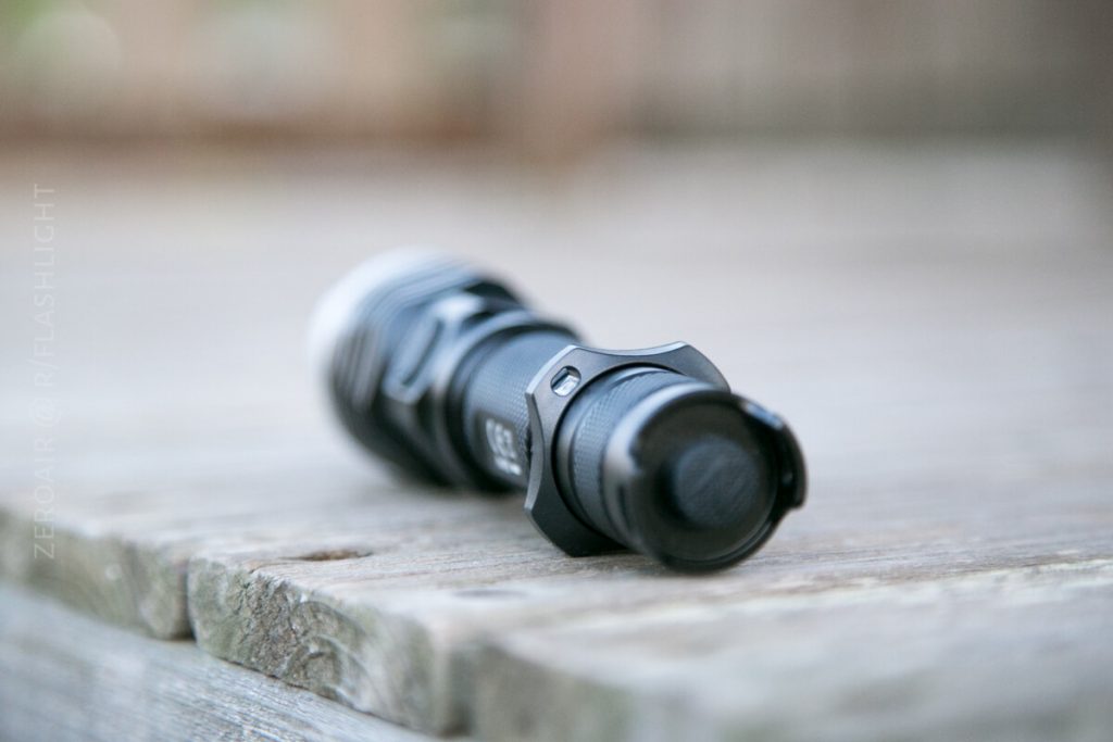 A black flashlight lies on its side on a wooden surface, with the focus on the tail switch. The background is blurred.