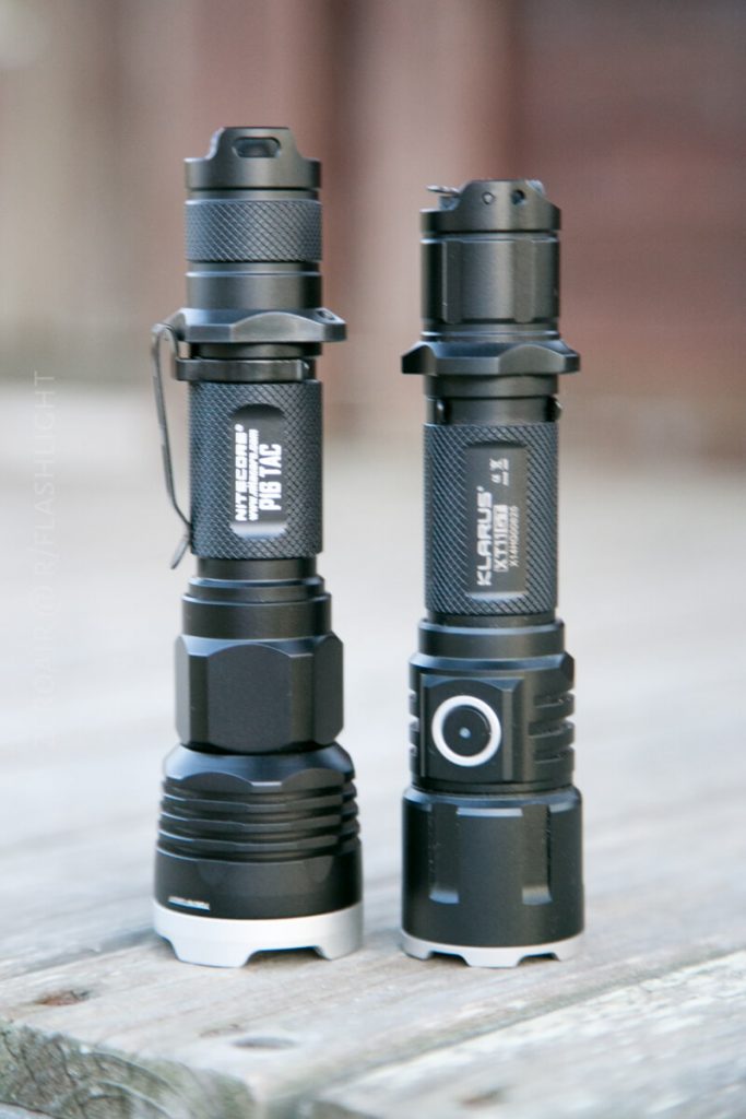 Two black tactical flashlights stand upright on a wooden surface. Both have textured grips and silver bases, with brand names and model numbers visible on their bodies. The background is blurred.