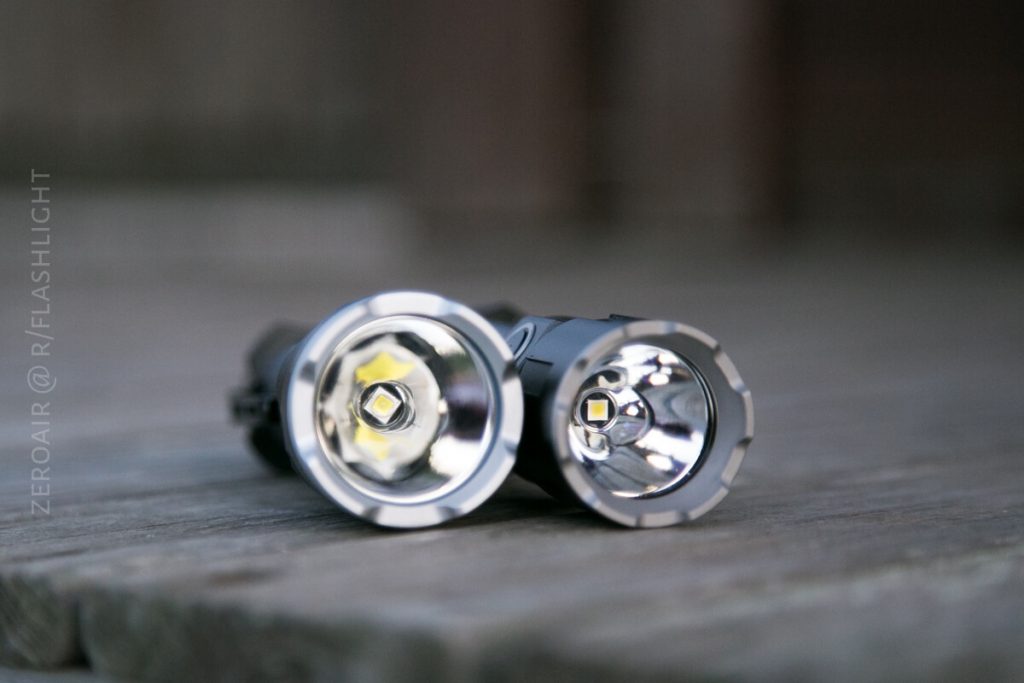 Two LED flashlights are placed side by side on a wooden surface, both facing the camera with their lenses and reflectors clearly visible. The image background is softly blurred.