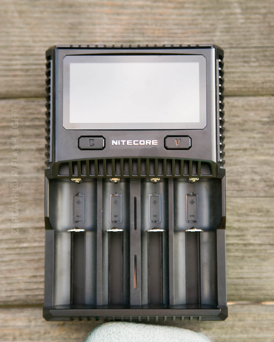 A Nitecore battery charger with a digital display and four battery slots is placed on a wooden surface. The charger is empty, and buttons labeled “C” and “V” are visible below the screen.