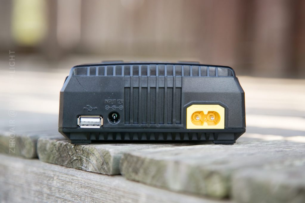 A close-up of the back side of a black electronic device on a wooden surface, showing a USB port, a round power input, and a yellow two-pin connector.