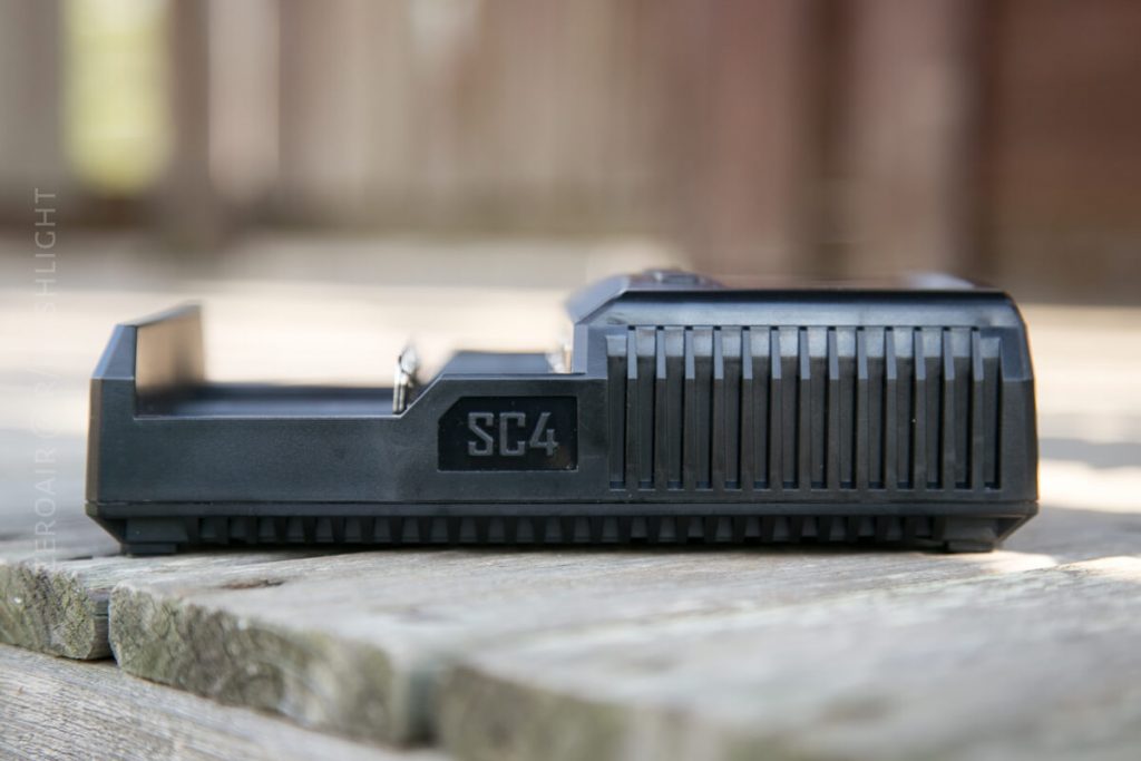 A black electronic device labeled SC4 sits on a wooden surface outdoors. The device features vertical ridges on the side and an open compartment, possibly for charging batteries. The background is blurred.