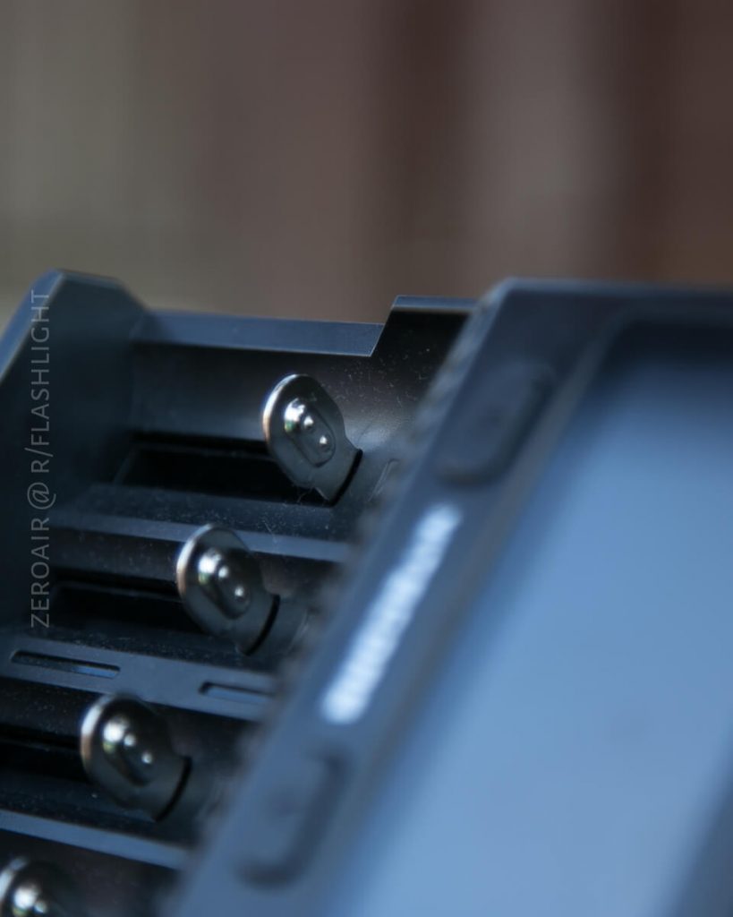 Close-up view of an empty battery charger with four slots, angled such that the metal contacts and part of the outer casing are visible. The background is out of focus.