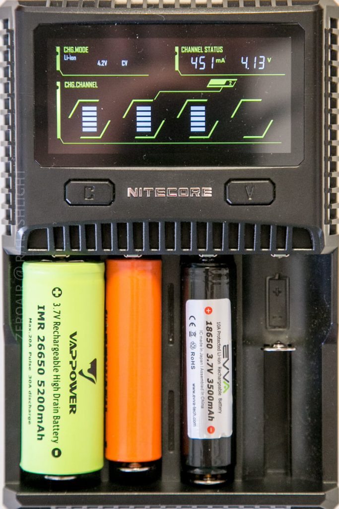 A Nitecore battery charger with two cylindrical batteries inserted, displaying charging information on a digital screen above, including voltage, charging current, and charge level bars for each battery slot.