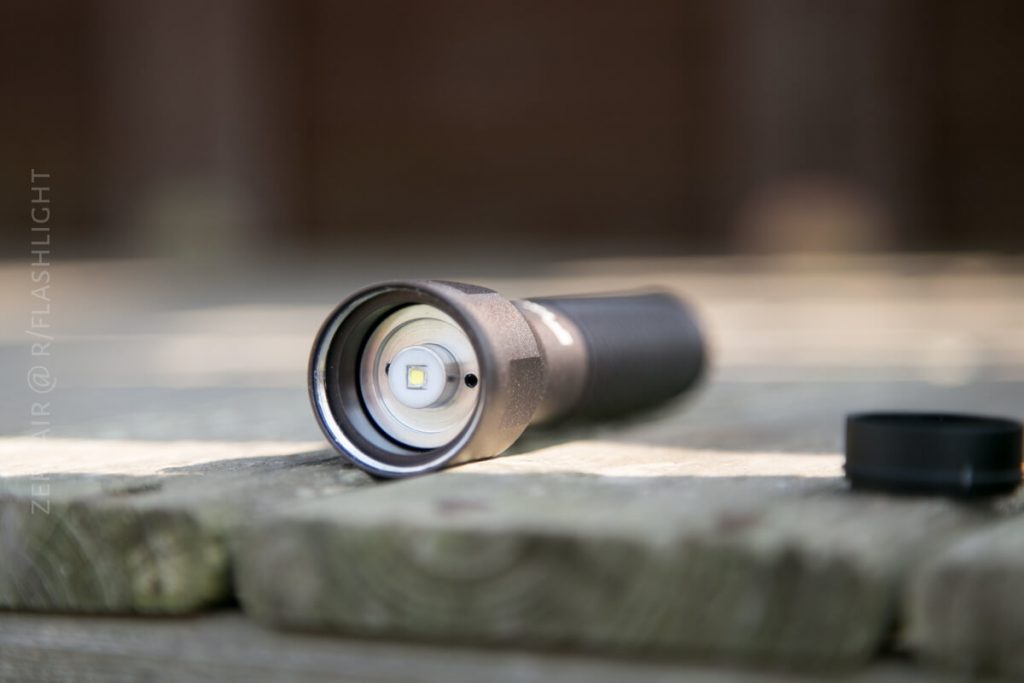 A black flashlight lies on a wooden surface with its lens and LED bulb facing the camera. The cap is removed and placed nearby. The background is blurred.