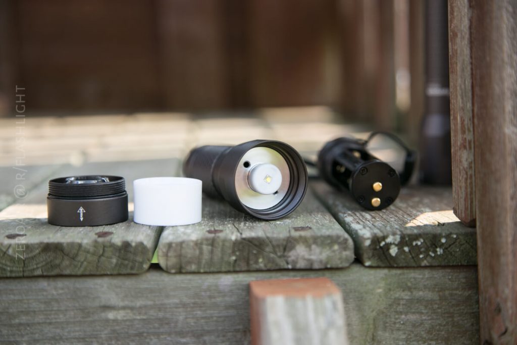 A disassembled black flashlight lies on a weathered wooden surface, with its body, battery compartment, and white plastic part separated and aligned next to one another.