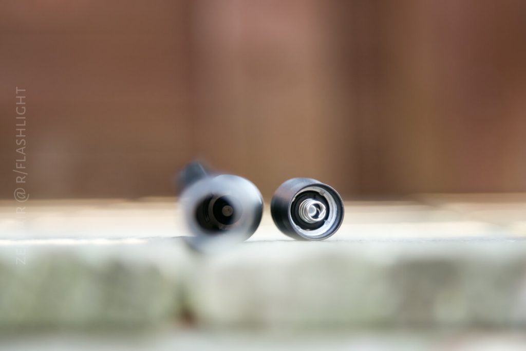 Two flashlight tubes are lying horizontally on a surface with their ends facing the camera, showing the battery contacts inside. The background is blurred.