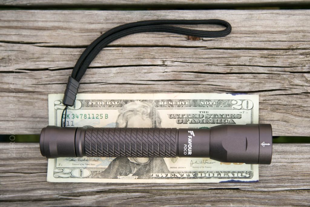 A black flashlight with a wrist strap is placed on top of a twenty-dollar bill on a weathered wooden surface. The flashlight is about the same length as the bill.