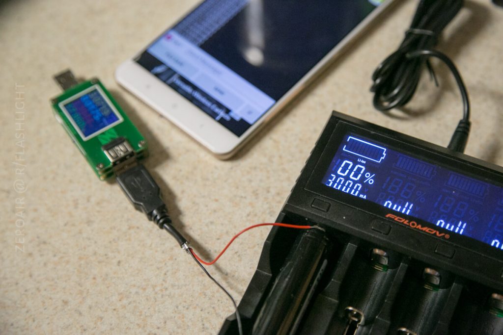 A battery charger with digital display is connected to a USB power meter and a white smartphone on a beige countertop. The charger shows battery and voltage information. A black USB cable connects the devices.