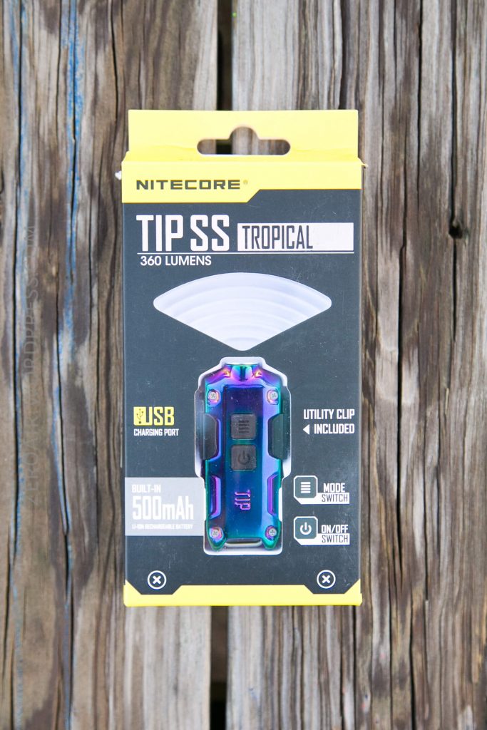 A Nitecore TIP SS Tropical 360 lumens flashlight in packaging, featuring a colorful metallic finish, USB charging port, utility clip, and 500mAh battery, displayed against a wooden background.
