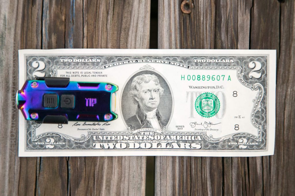 A multicolored metal money clip is attached to the left side of a two-dollar bill featuring Thomas Jefferson, placed on a wooden surface.