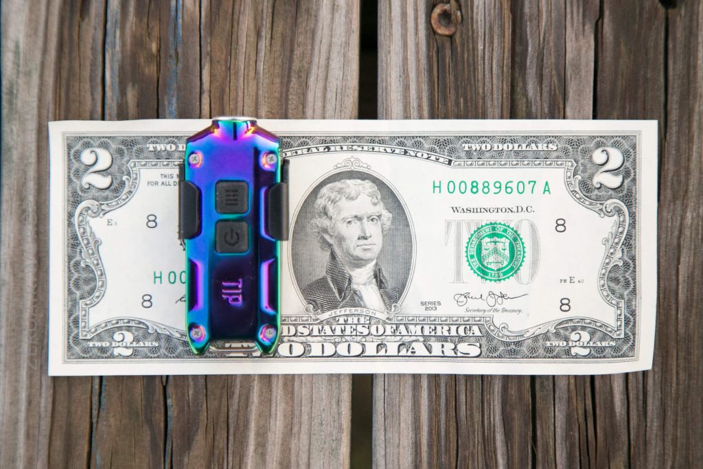 A $2 bill is clipped to a wooden surface with a metallic, multi-colored money clip or tool. The bill features Thomas Jefferson’s portrait and a green Treasury seal.