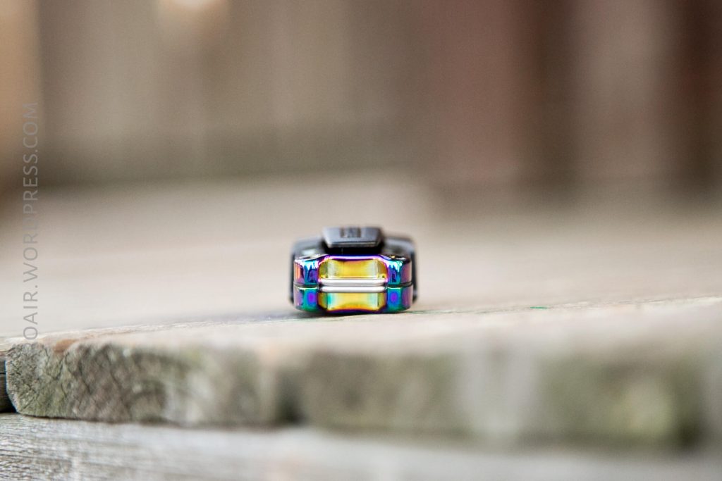 Close-up of a small, metallic object with a rainbow sheen placed on a wooden surface. The background is blurred, and the object is positioned centrally in the image.