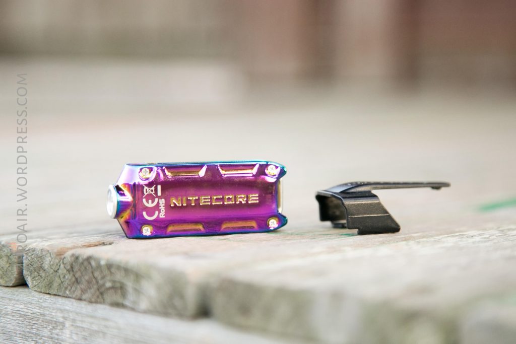 A small, iridescent Nitecore keychain flashlight and a black plastic clip are placed on a weathered wooden surface. The flashlight branding and model are visible.