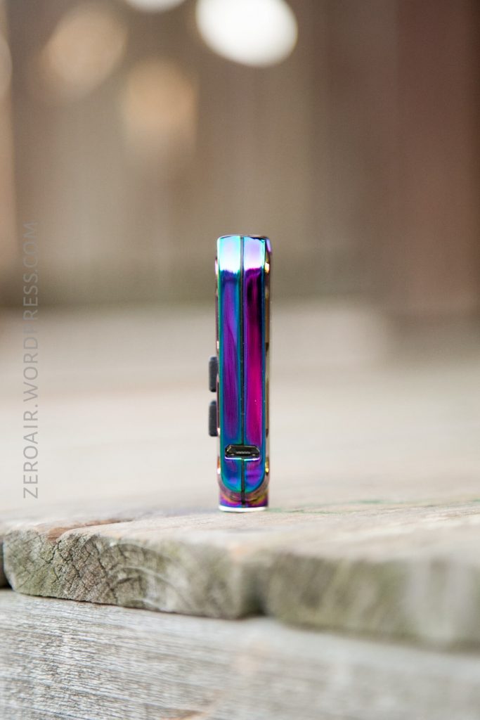 A small, metallic, iridescent device with buttons is standing upright on a wooden surface with a blurred background. The device has a reflective finish showing shades of blue, green, and purple.