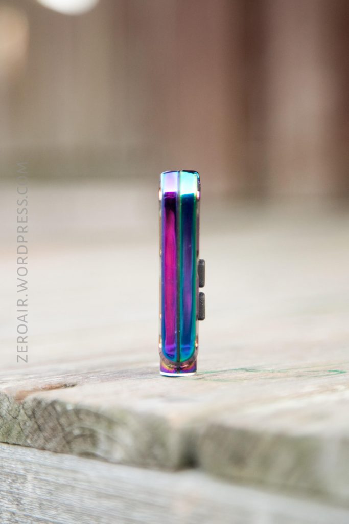 A small, iridescent metallic object with two black buttons stands upright on a wooden surface. The background is blurred. The text ZEROAIR.WORDPRESS.COM is faintly visible along the left side.