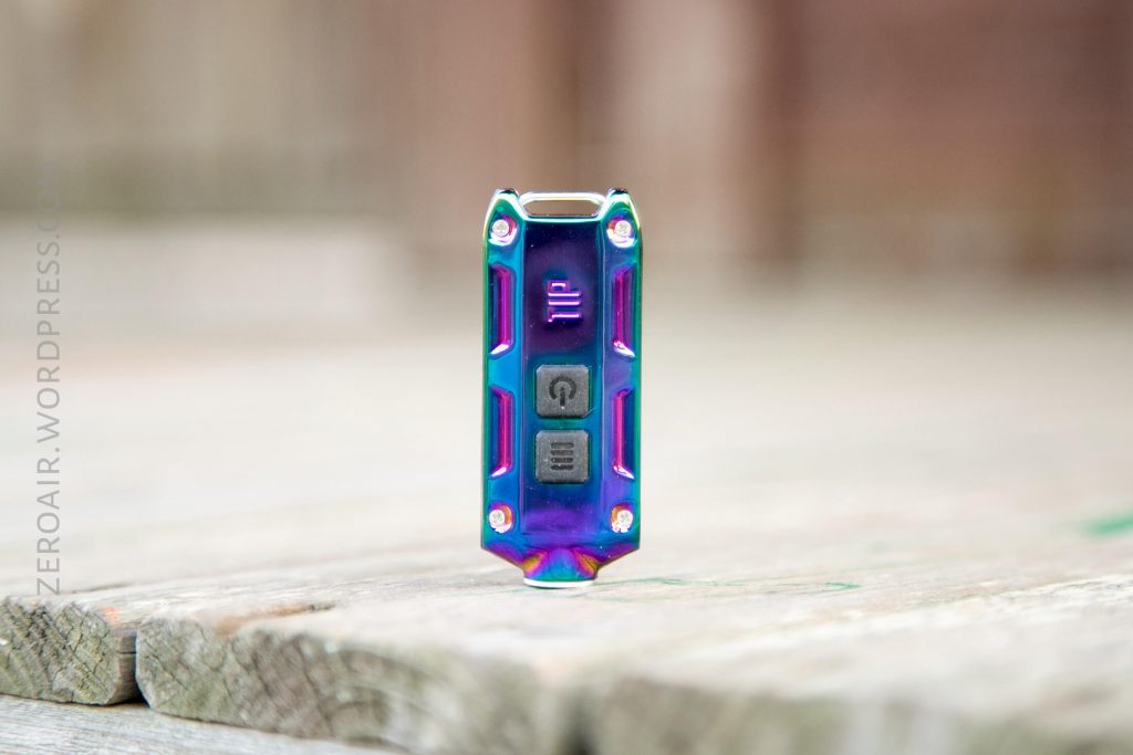 A small, metallic, rainbow-colored device with two black buttons and visible screws stands upright on a weathered wooden surface. The background is blurred.