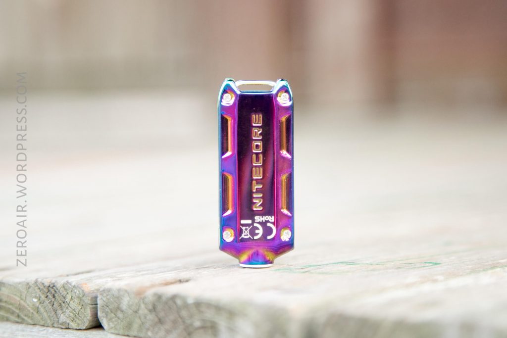 A small, metallic rectangular object with a shiny, iridescent finish and the word NITECORE printed on the front, standing upright on a weathered wooden surface.