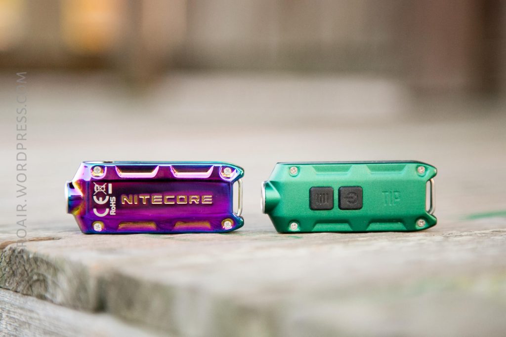 Two small Nitecore keychain flashlights rest on a wooden surface; one is metallic rainbow-colored showing the front, and the other is matte green showing the back with two buttons and a charging port.