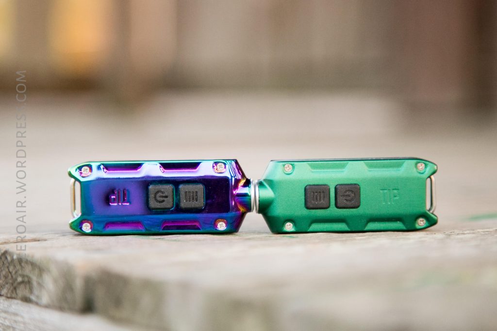 Two compact flashlights lie side by side on a wooden surface. The flashlight on the left has a metallic, iridescent finish, while the one on the right is matte green. Both have black buttons and labeled switches.