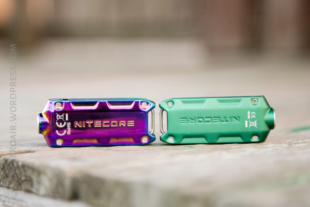 Two Nitecore keychain flashlights rest on a wooden surface, one with a metallic rainbow finish and the other with a matte green finish, both displaying the brand name and certification markings.