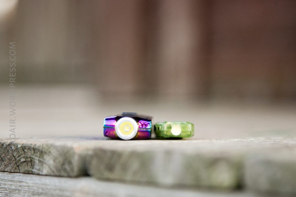 Two small LED flashlights lie side by side on a wooden surface, with one flashlight being metallic purple and the other translucent green. The image is focused on the lights, with a blurred background.