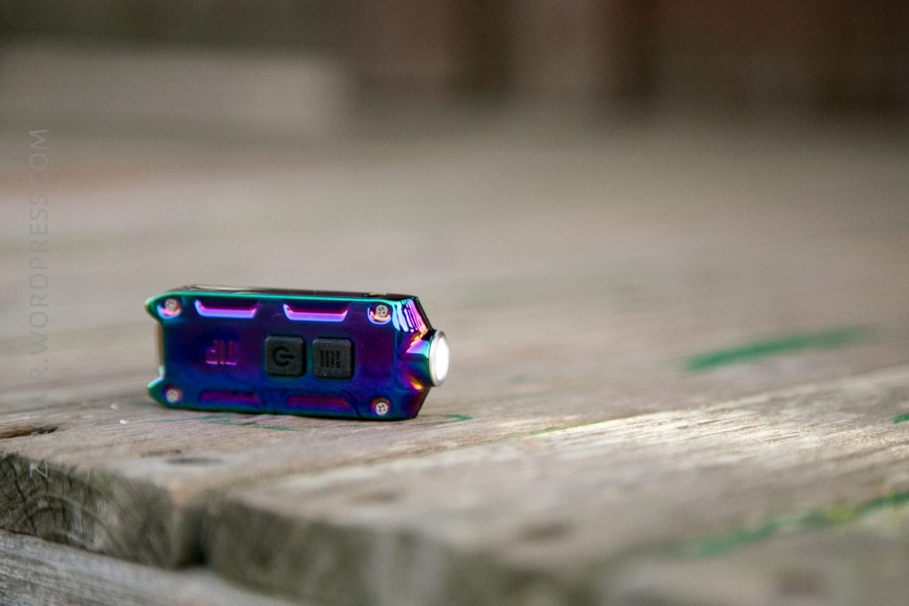 A small, iridescent flashlight with visible buttons lies on a weathered wooden surface, slightly out of focus in the background.