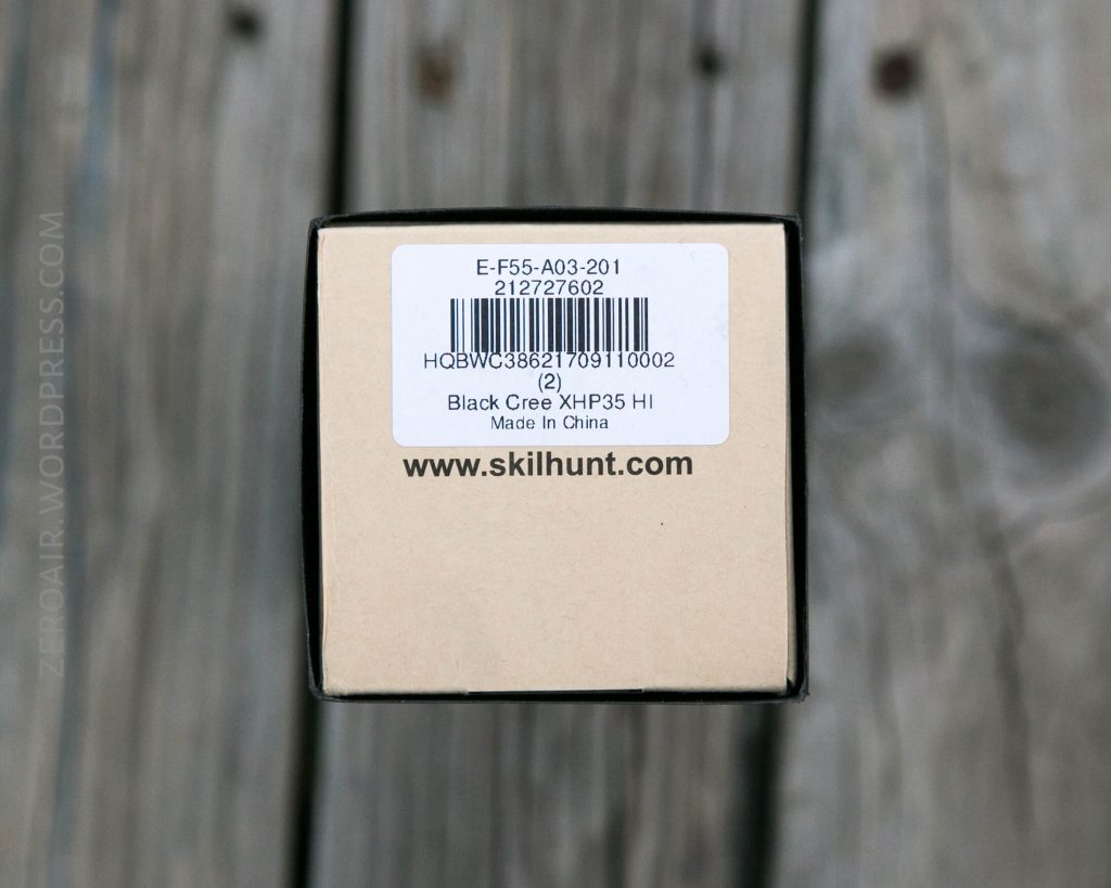 A cardboard box with a white barcode label on top. The label reads Black Cree XHP35 HI Made in China and includes item numbers and the website www.skilhunt.com. The box is on a wooden surface.