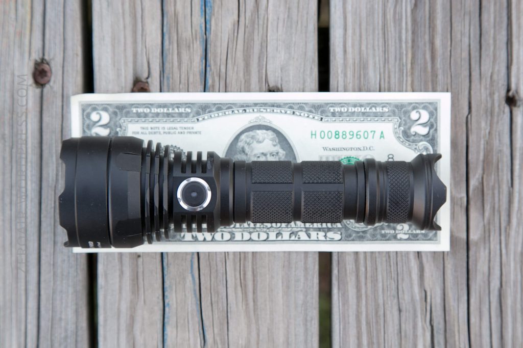 A black flashlight lies horizontally on top of a two-dollar bill, both placed on a wooden surface. The flashlight is slightly longer than the bill.