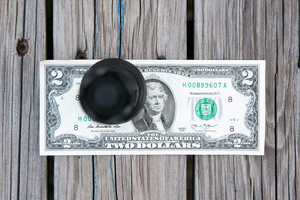 A glass upside down is placed on top of a two-dollar bill on a wooden surface with visible grain and gaps between the planks.