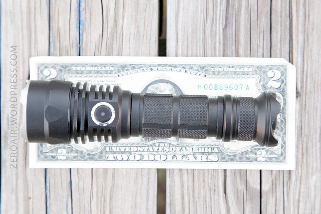 A black flashlight is placed horizontally on top of a two-dollar bill, which sits on a wooden surface. The flashlight is centered, and the bill shows part of the serial number and US Treasury details.