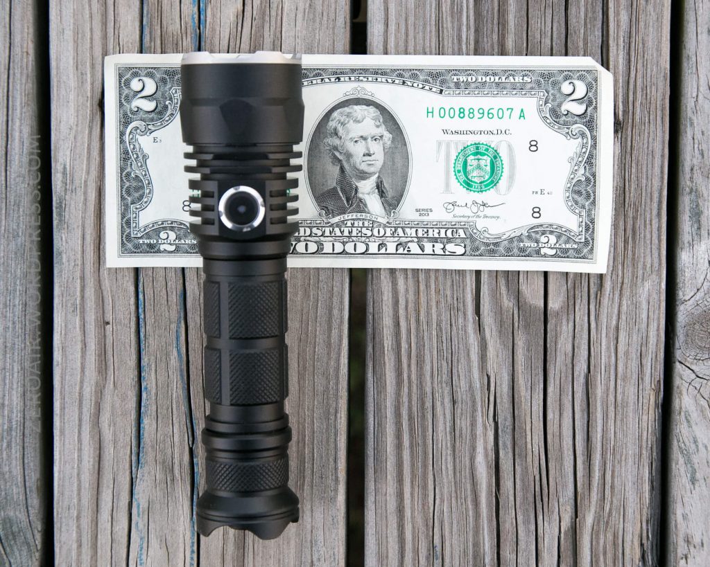 A black flashlight is placed upright in front of a U.S. two-dollar bill, both resting on a weathered wooden surface. The flashlight is nearly as tall as the bill.