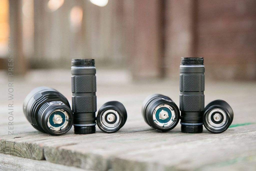 Three black flashlights are disassembled and arranged on a wooden surface, each with the head, battery tube, and tail cap separated and standing upright in a row. The background is slightly blurred.