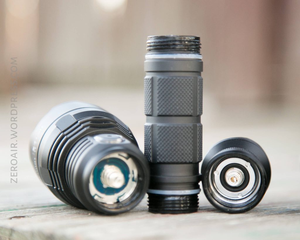 A close-up of a disassembled black flashlight showing the head, battery tube, and tailcap placed on a wooden surface. The interior threads and metal contacts are visible.