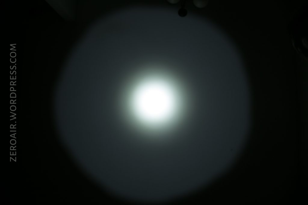 A flashlight beam projects a circular white light with a bright center onto a dark background. The beam has a defined edge and a softer halo surrounding the middle. ZEROAIR.WORDPRESS.COM is written vertically on the left.