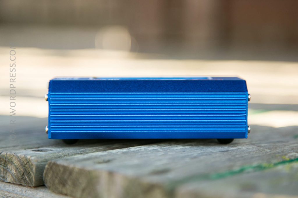 A small blue rectangular metal box with ridged sides is placed on a wooden surface outdoors. The box has four small feet and no visible labels or features on its front side.