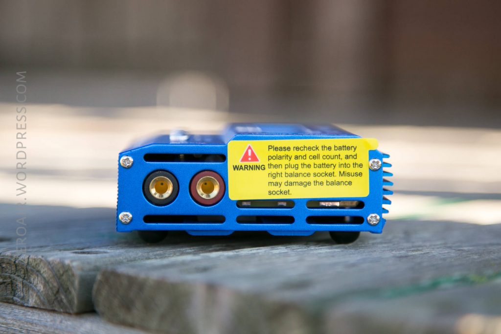 A close-up of a blue electronic device with two circular ports and a yellow caution label that warns about checking battery polarity and using the correct socket to avoid damage. The device is placed on a wooden surface.