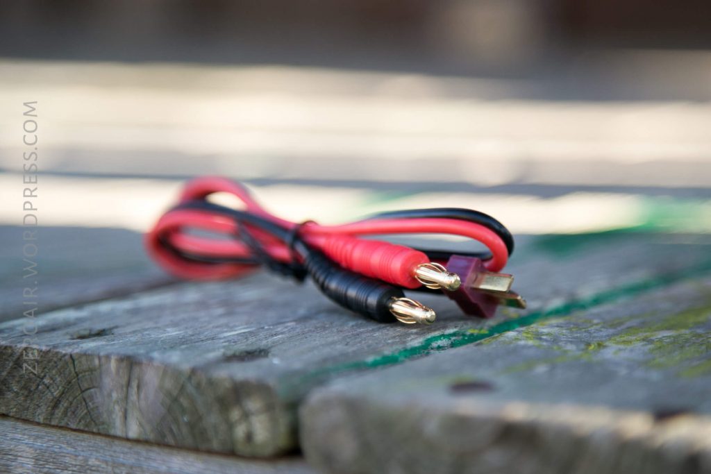A red and black electrical cable with gold-plated banana plugs lies coiled on a weathered wooden surface. The background is blurred.