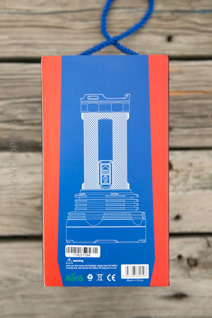 A blue and red product box with a white outline illustration of a flashlight on the front, various certification icons, barcodes, and a recycling symbol are visible at the bottom. The box is placed on a wooden surface.