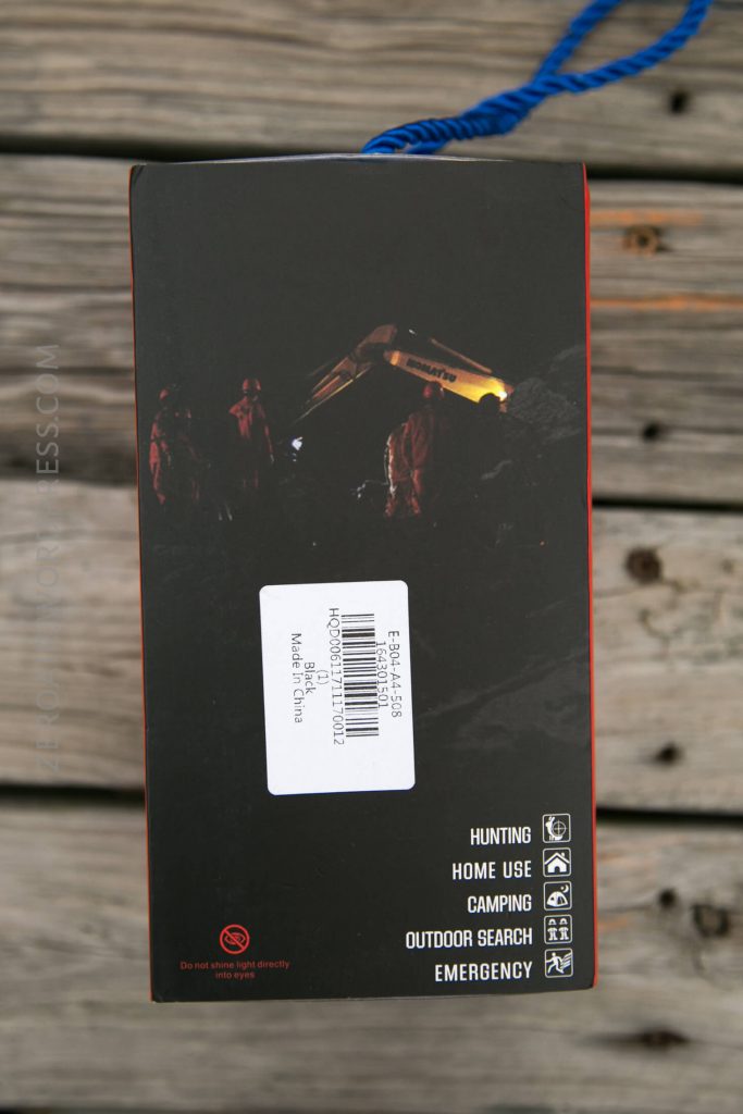 A product box sits on wooden boards. The box shows a nighttime scene with people near an excavator. Text at the bottom lists uses: hunting, home use, camping, outdoor search, and emergency. A barcode label is on the box.