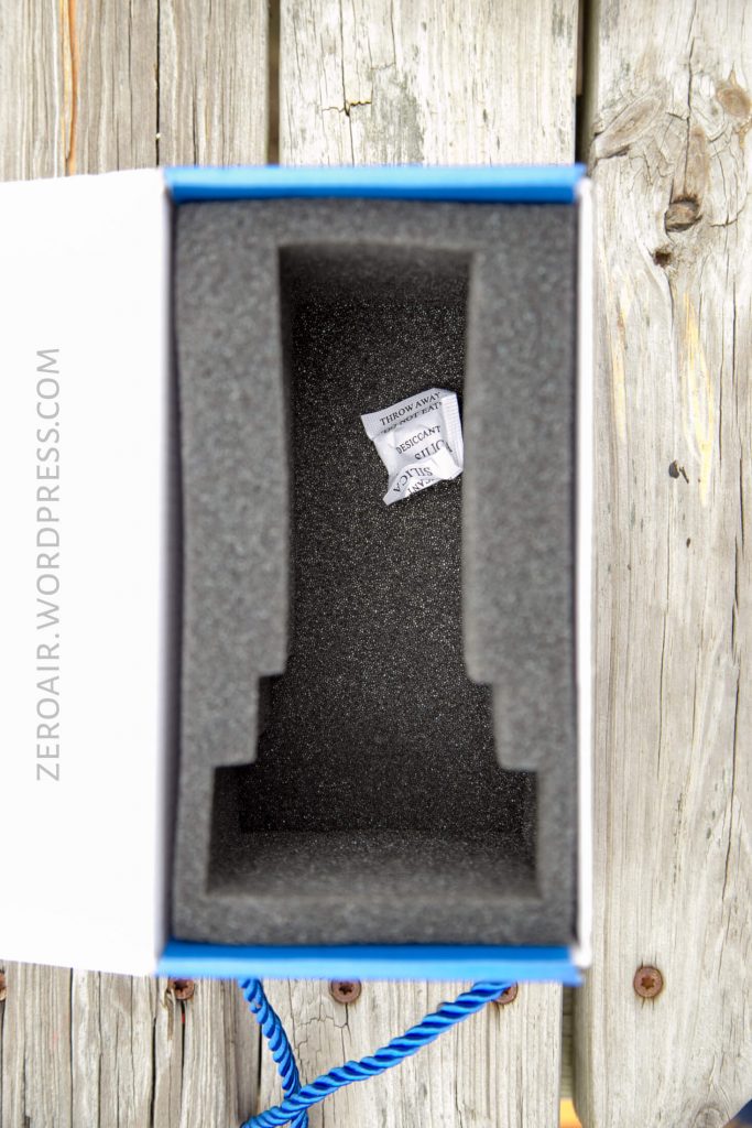 A rectangular foam insert with a small packet labeled “SILICA GEL DO NOT EAT” sits inside an open box. The box is placed on a weathered wooden surface. The side reads “ZEROAIR.WORDPRESS.COM.”