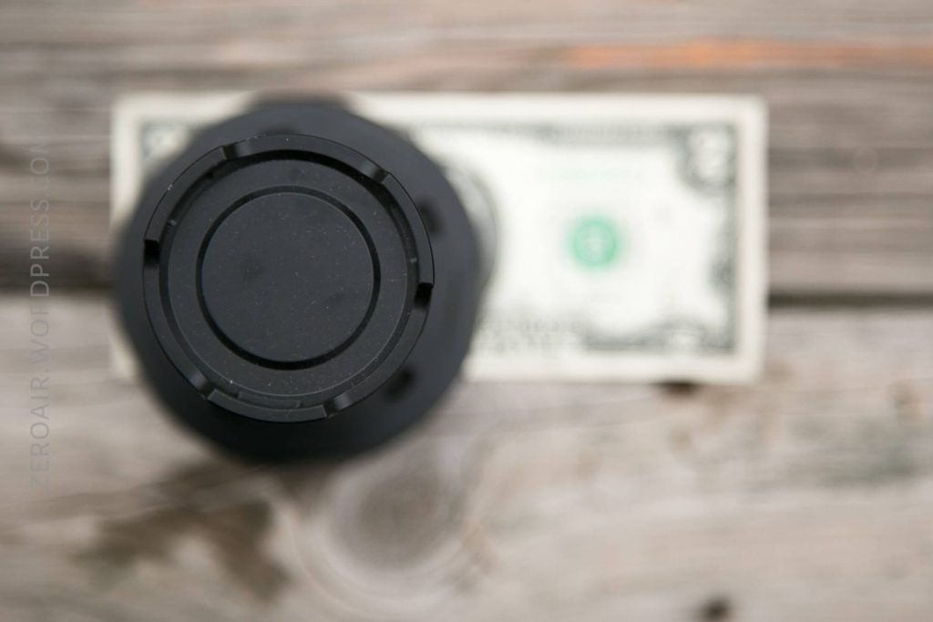 A black cylindrical object is centered in the foreground, viewed from above, with a blurred U.S. one-dollar bill placed on a wooden surface in the background.