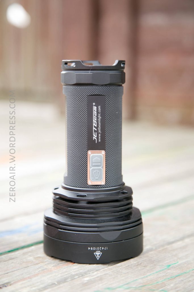 A black JETBeam flashlight with a textured grip stands upright on a wooden surface. The power button and branding are visible on the flashlight’s body. Background is out of focus.