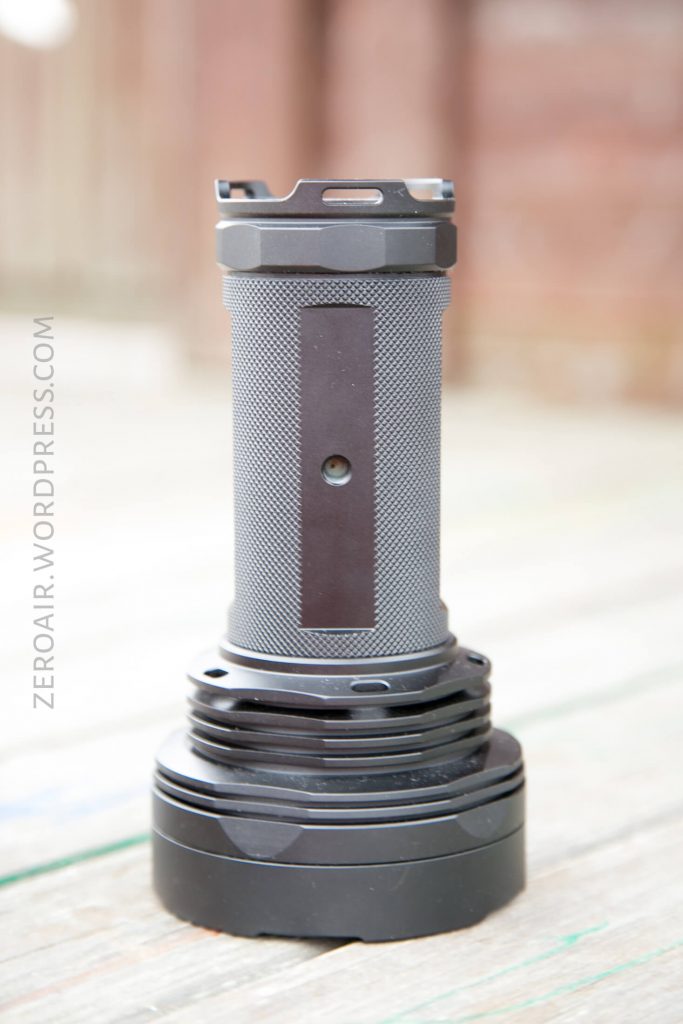 A large, black, cylindrical flashlight with a textured grip stands upright on a wooden surface. The background is blurred. The text ZEROAIR.WORDPRESS.COM is faintly visible along the left side.