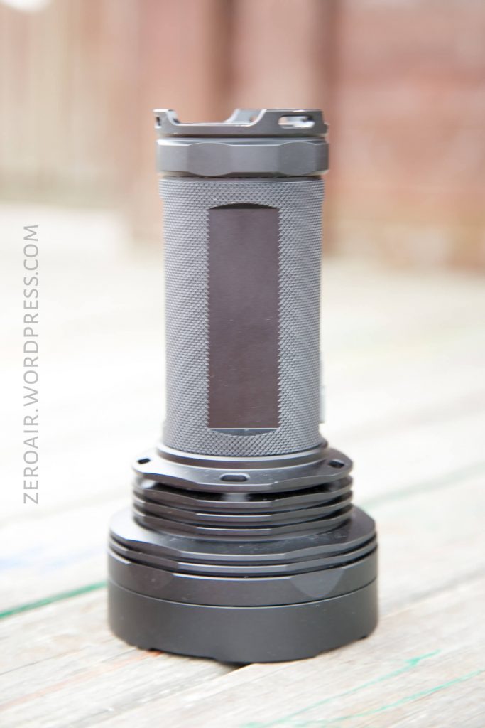 A large, cylindrical flashlight with a textured grip stands upright on a wooden surface. The background is blurred. The flashlight is mostly gray and black. The words “ZEROAIR.WORDPRESS.COM” appear along the left side.