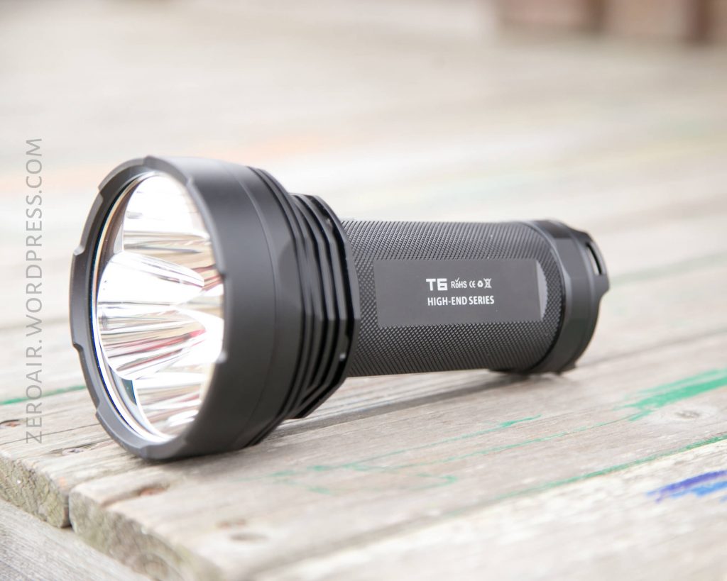 A black handheld flashlight labeled TE High-End Series lies on a weathered wooden surface, with its lens facing left. The handle has a textured grip and reflective elements are visible inside the head.