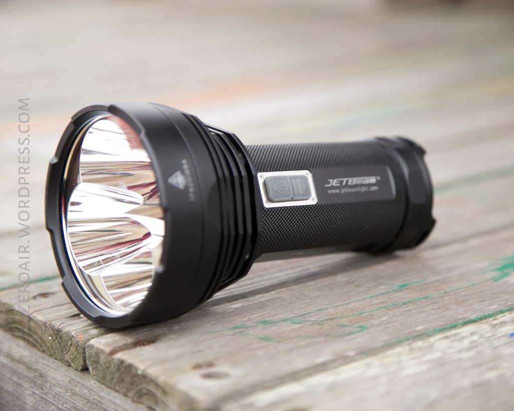 A black JETBeam flashlight with a textured grip and three reflectors is lying on its side on a weathered wooden surface. The flashlight is positioned at an angle, with branding and website visible.