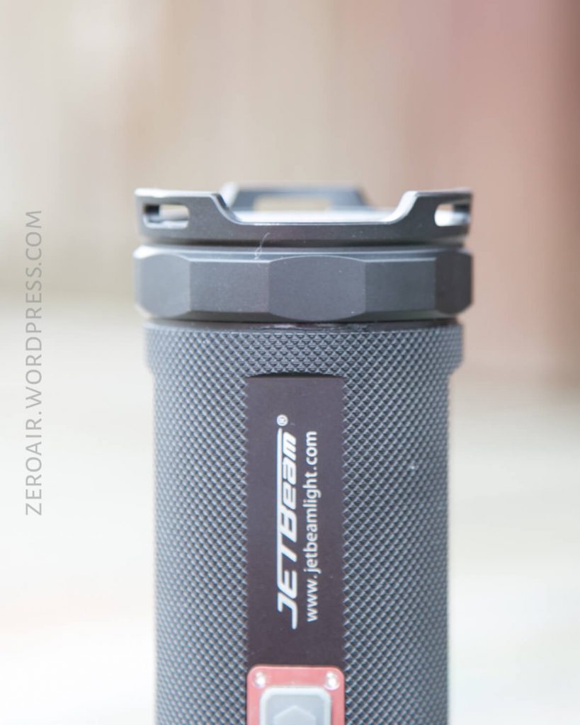 A close-up of a black JETBeam flashlight standing upright, showing its textured grip, metal bezel, and brand label. The background is blurred. The text “ZEROAIR.WORDPRESS.COM” is visible along the left side.