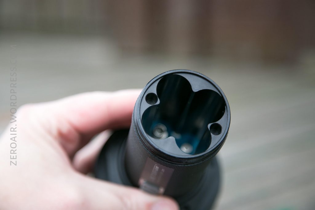 A close-up of a hand holding a cylindrical electronic device with a rounded, black three-slot opening at the top, showing two visible metal contacts inside. The background is blurred.