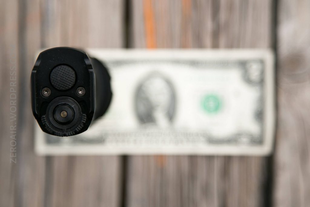 A close-up, top-down view of the barrel of a black handgun pointed downward at a U.S. one-dollar bill placed on a wooden surface. The image is slightly out of focus around the bill.